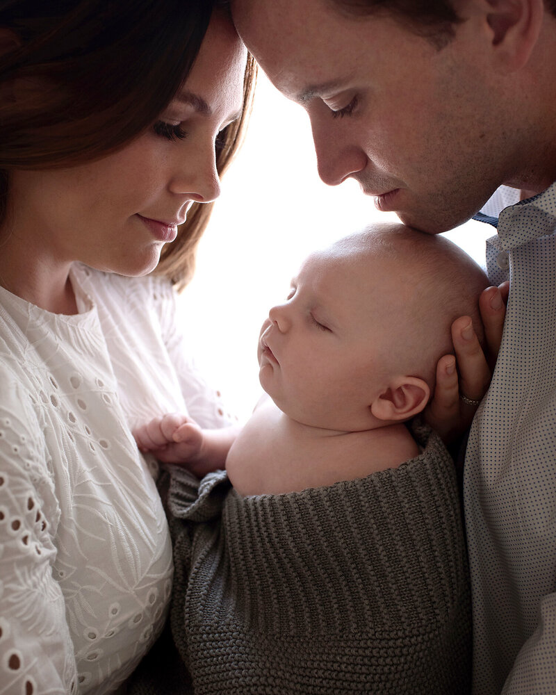 Parents standing in front of a window cradling their newborn son between them at a lifestyle newborn session with Susan Baraban Photography.