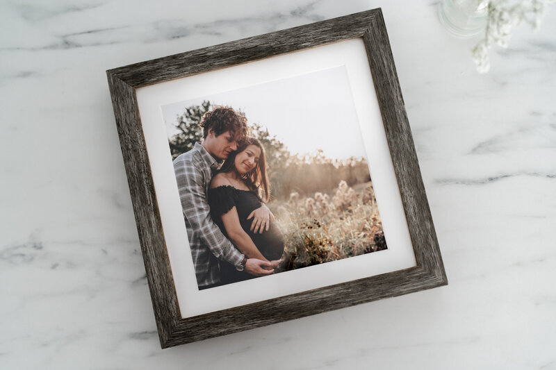 A framed maternity photo of a couple standing in a field at sunset. The man is embracing the woman from behind as they both smile and look down at her belly.