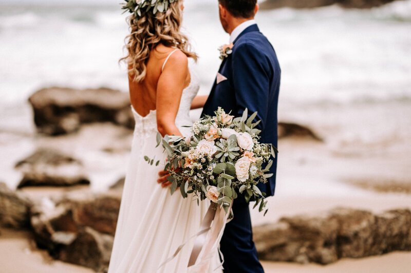 A bride and groom stood on the beach in Cornwall looking at the sea. The bride has flowers in her hair and carrying a large bouquet of peach roses, foliage and succulents