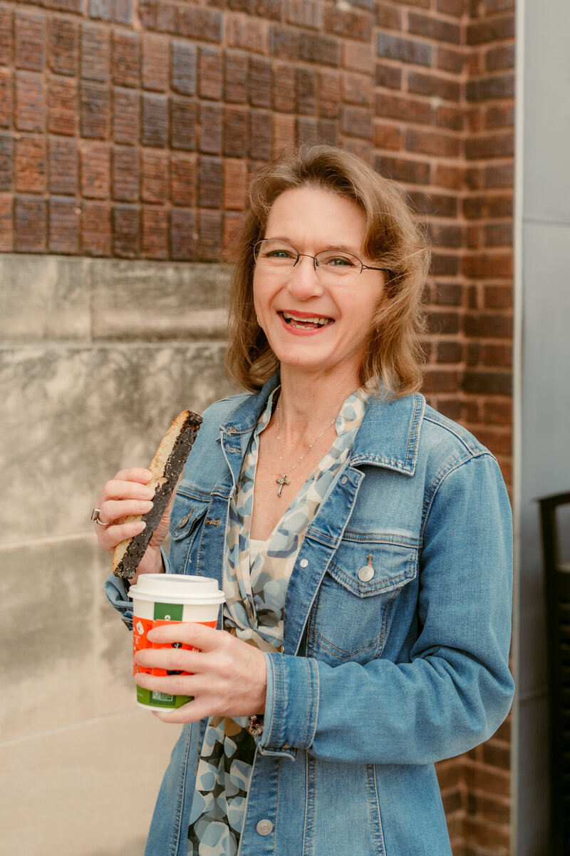 A woman smiling outdoors while holding coffee and a pastry during a natural-light online dating photo session, photographed by Shannon Kathleen Photography to capture her warm, approachable personality.