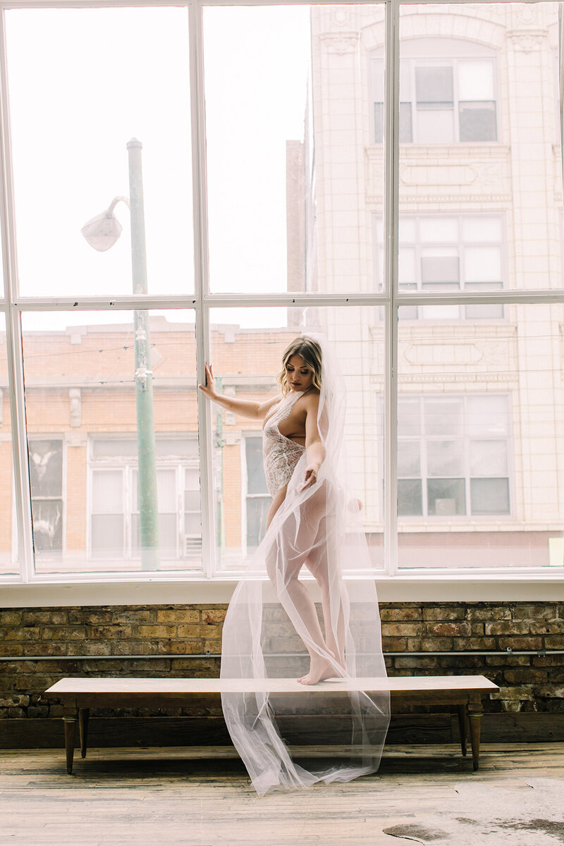 Bride poses in window while wearing white lingerie and wedding veil.
