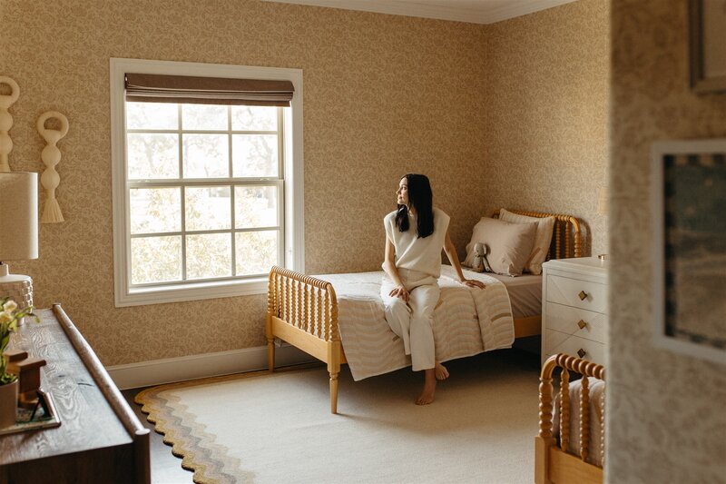 Hope Danyla sitting on a bed in a warm, coastal bedroom, designed by Hope Miriam Designs in DFW