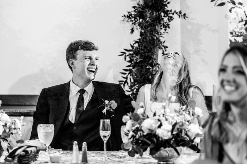 Spring wedding at WinMock at Kinderton, designed by Charming Carolina — newlyweds seated at the head table, surrounded by floral centerpieces and taper candles, smiling during reception toasts.