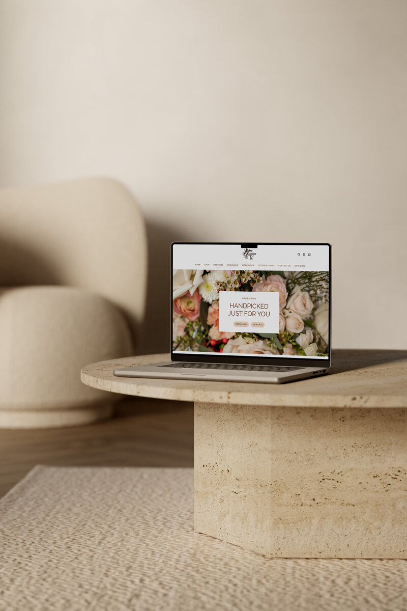 An open laptop sits on a stone coffee table and displays a still image of the website homepage for a luxury wedding florist called Alpine Designs & Landscaping