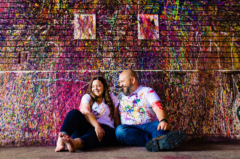 Engaged couple sitting in a spatter paint studio coverred in colorful paint laughing in Toledo Ohio