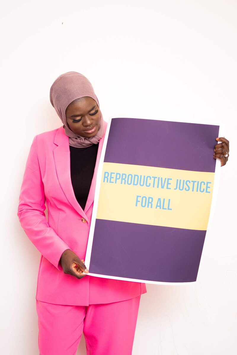 Professional grant writer at Amikha Consulting holding a sign that says "reproductive Justice for all". She is wearing a taupe headwrap, and wearing a pink blazer with a black shirt underneath.