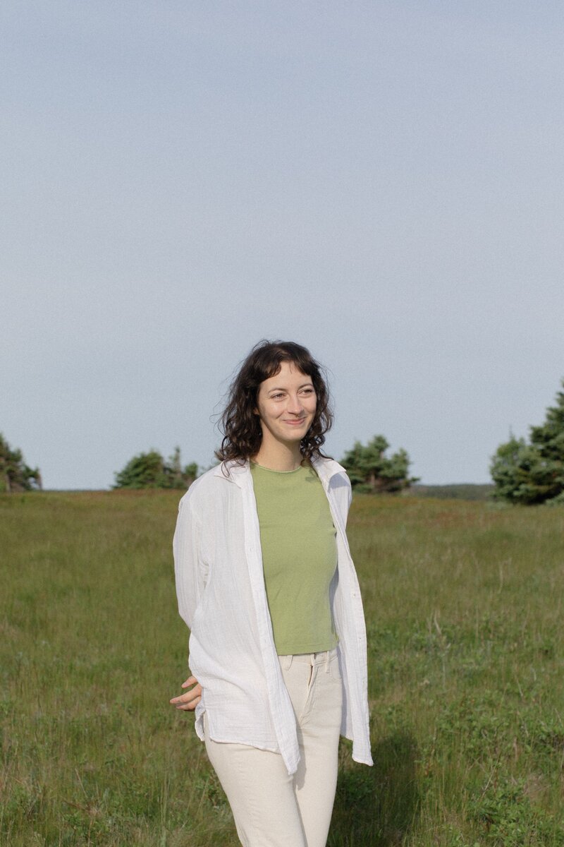 Heather smiles serenely in a meadow. They are a white person with curly brown hair, wearing a green top and white pants.