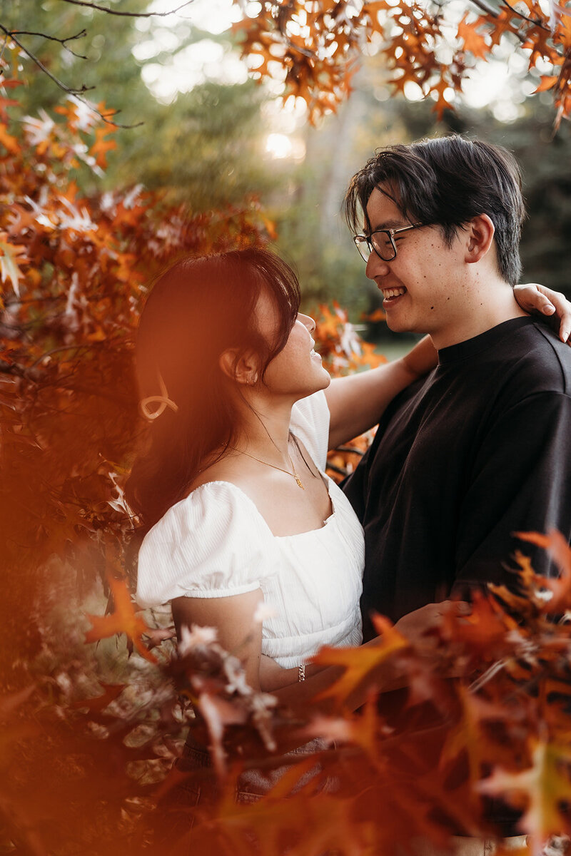 Mid shot of engaged couple looking into each other eyes. Beautiful golden autumn leaves in the background and foreground of the photo. Photography by Acacia and Grace.