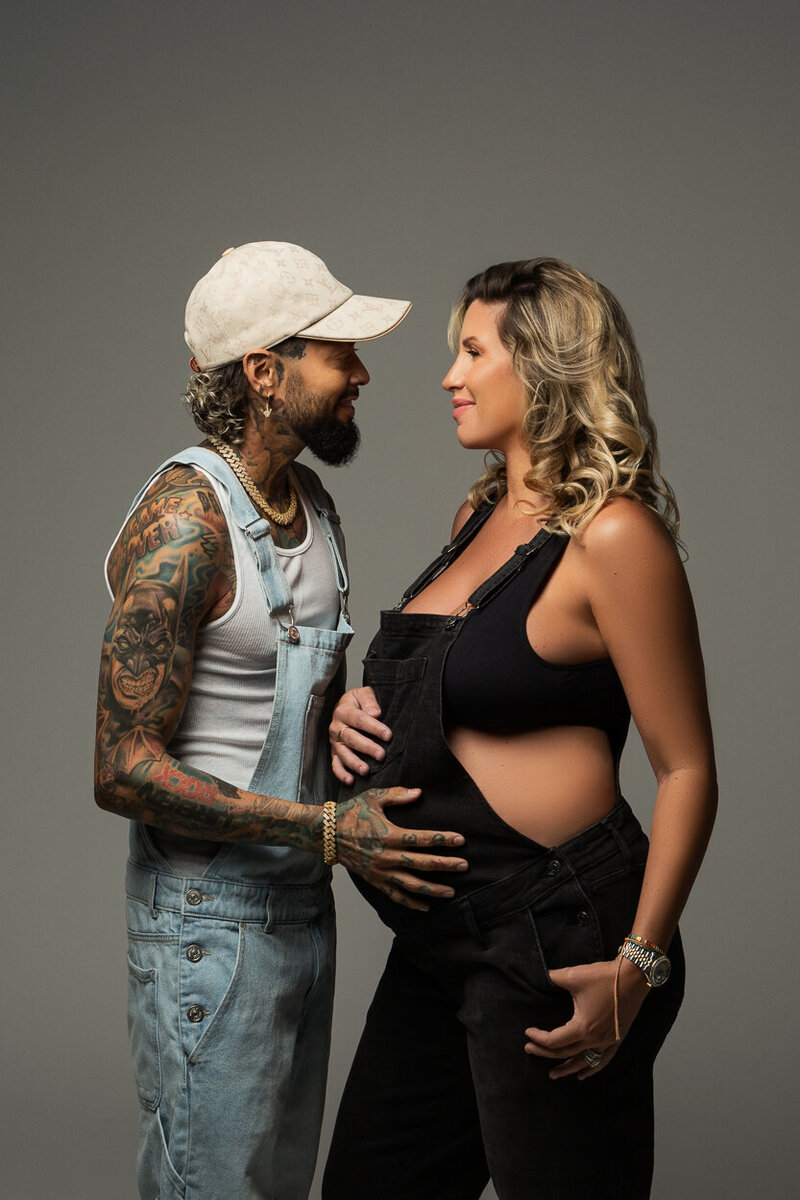 Charlotte couple wearing overalls gazing into each others eyes both holding mom to be's pregnant belly. Posing in the Insley Photo studio in front of a grey background. Dad is hispanic and mom is caucasian