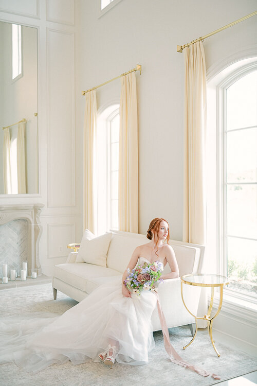 Elegant bridal portrait of bride sitting on white couch holding pastel floral bouquet at hillside estate wedding in Dallas