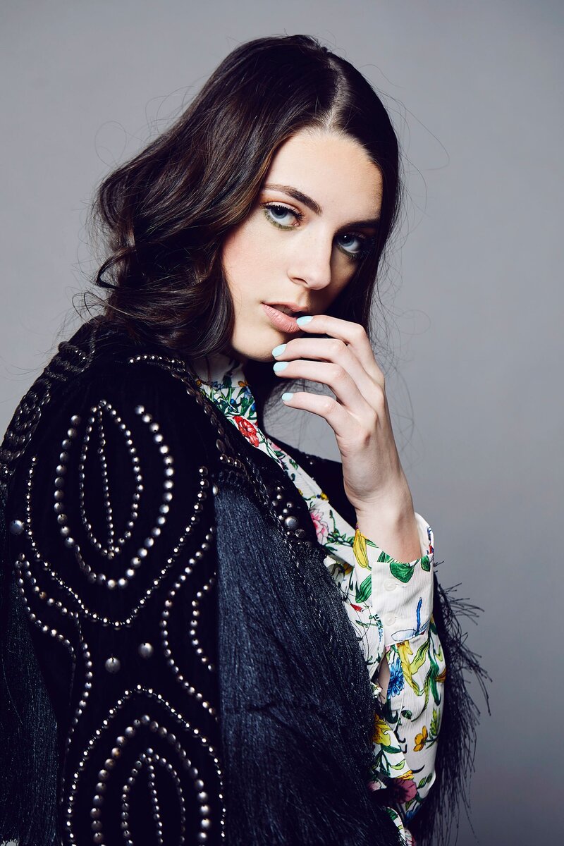 High-fashion close-up portrait of a teen girl with sleek dark hair wearing a detailed black beaded jacket, softly touching her lips and gazing at the camera.