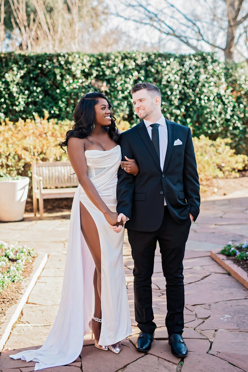 A couple locks arms and smiles at each other in a green sunny pathway. The woman wears a white silk gown with a leg slit.