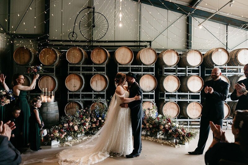 A bride and groom hugging at a floral alter on the ground in front of a wall of wine barrels.