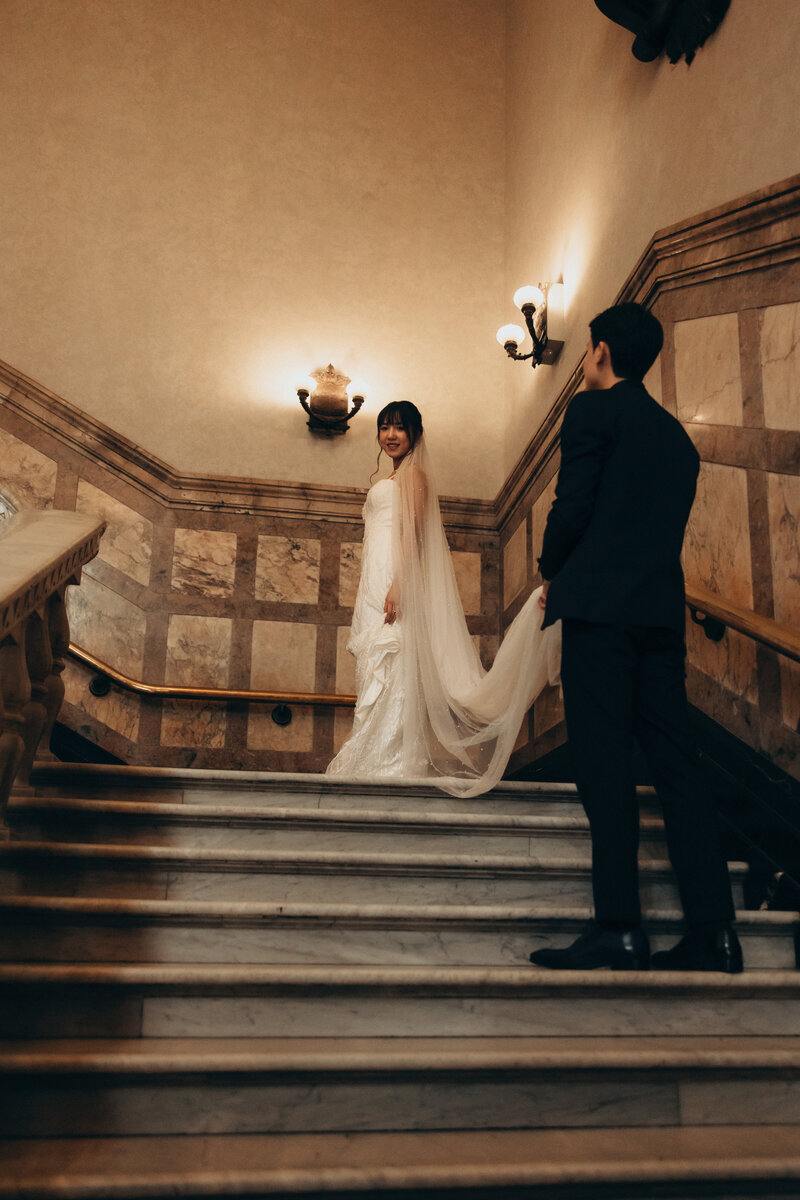 Bride and groom during an intimate elopement, with the bride smiling back while standing on a grand staircase, captured in an elegant heritage building.