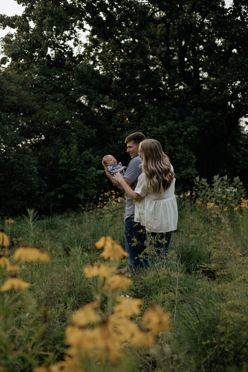 A family of three poses in a field of yellow flowers. Mom and Dad's back is turned away from the camera as they look at their newborn son that Dad is holding.