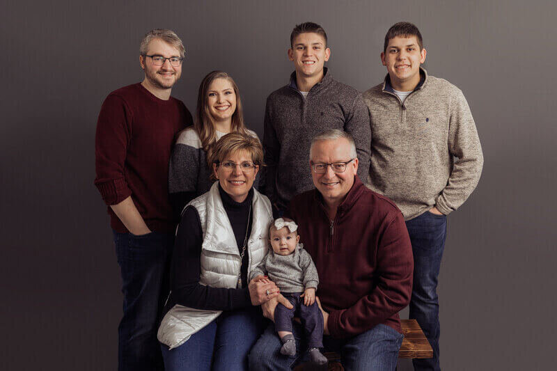 Studio family portrait with grandparents holding a baby and surrounded by their adult children, all smiling against a neutral backdrop.