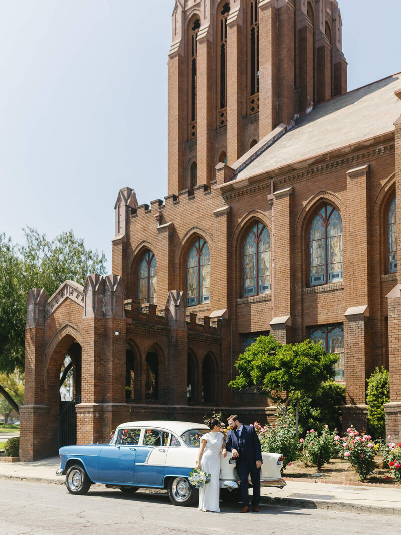 wide shot of bride and groom in front of church with classic blue car