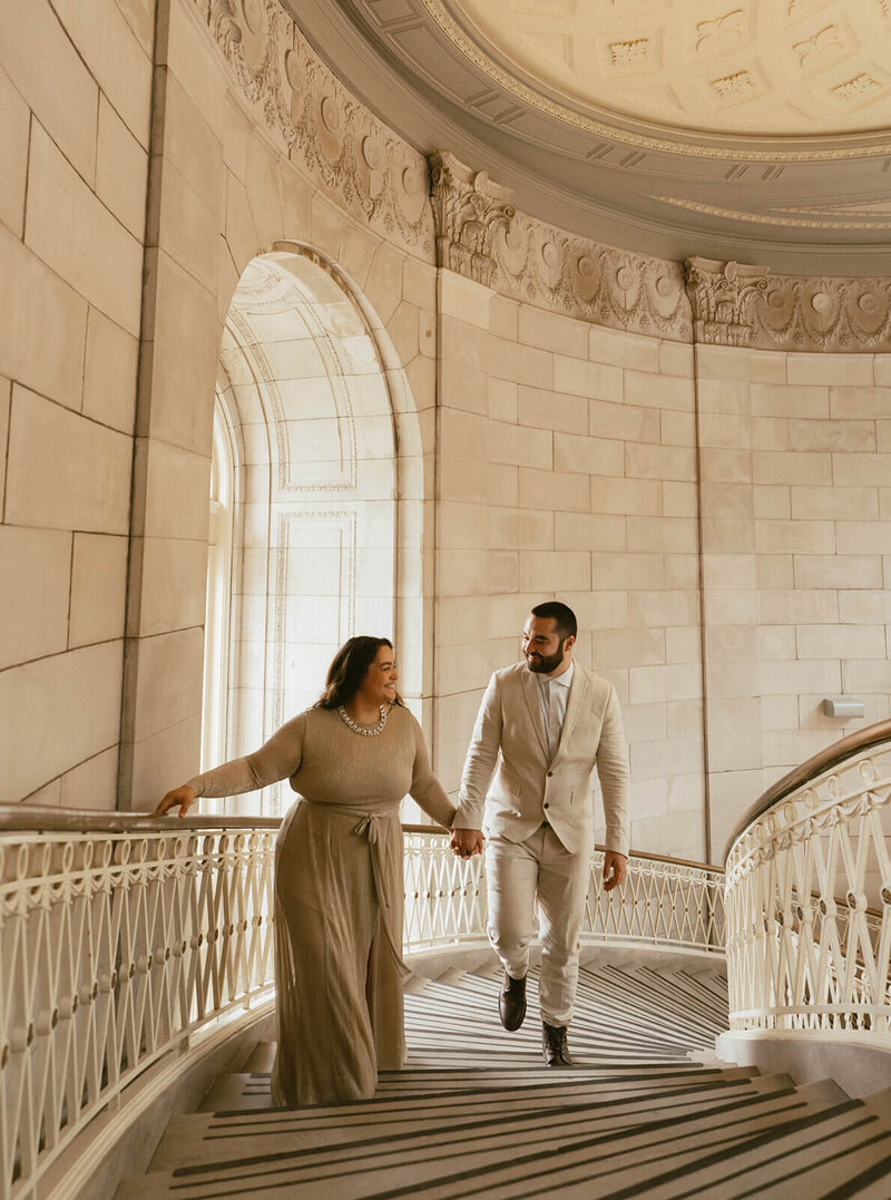 The groom and bride are walking up the stairs, holding hands at the City Hall