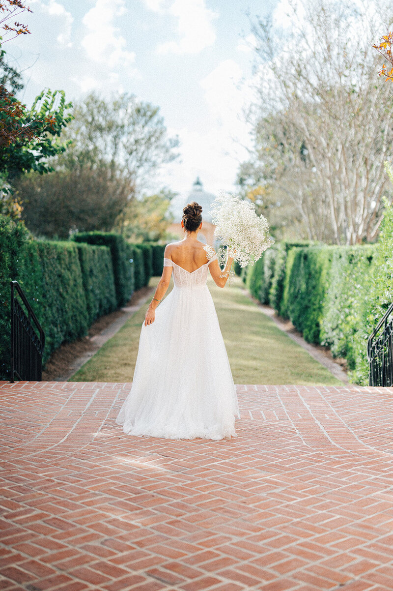 The photograph captures a bride from behind as she walks down a brick pathway lined with neatly trimmed hedges. She is wearing a flowing white gown with an open back and is holding a bouquet of Baby's Breath over her shoulder, creating a whimsical trail behind her.