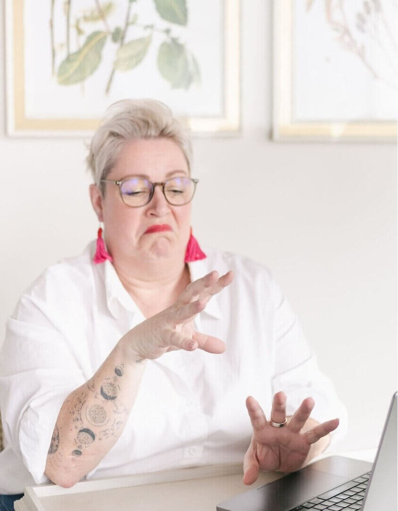 Pippa from Disobedient Business® Co. wearing glasses, bright pink earrings, and a white shirt, makes a humorous mid-conversation, disgusted expression while gesturing with both hands towards a laptop in a light, modern room.