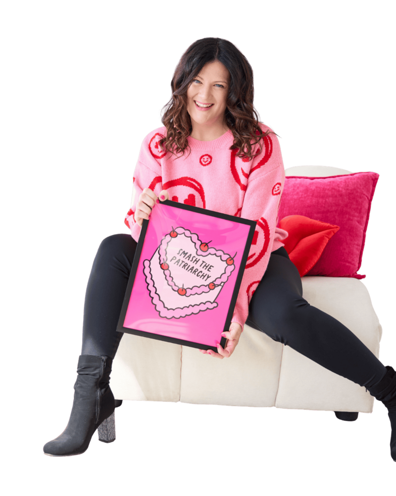 Emily sitting on a soft white chair, in a pink shirt, holding a brand illustration of a cake that says "Smash the Patriarchy."