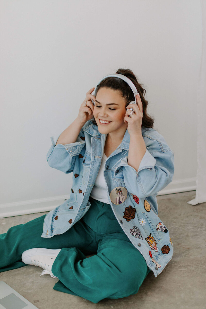 Woman with Dark hair with teal pants, jean jacket, white headphones sitting on floor