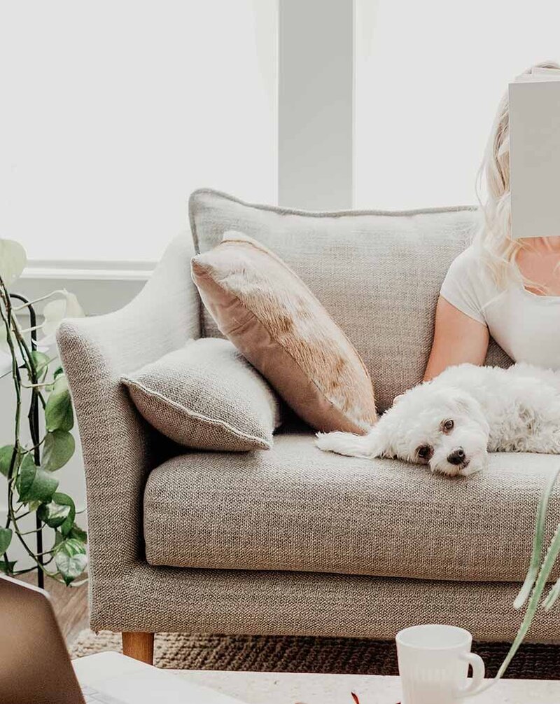 Young woman reading a book on a cozy couch with a white Maltese dog and a laptop on the coffee table, representing compassion in Xanthe Bookkeeping’s business values.