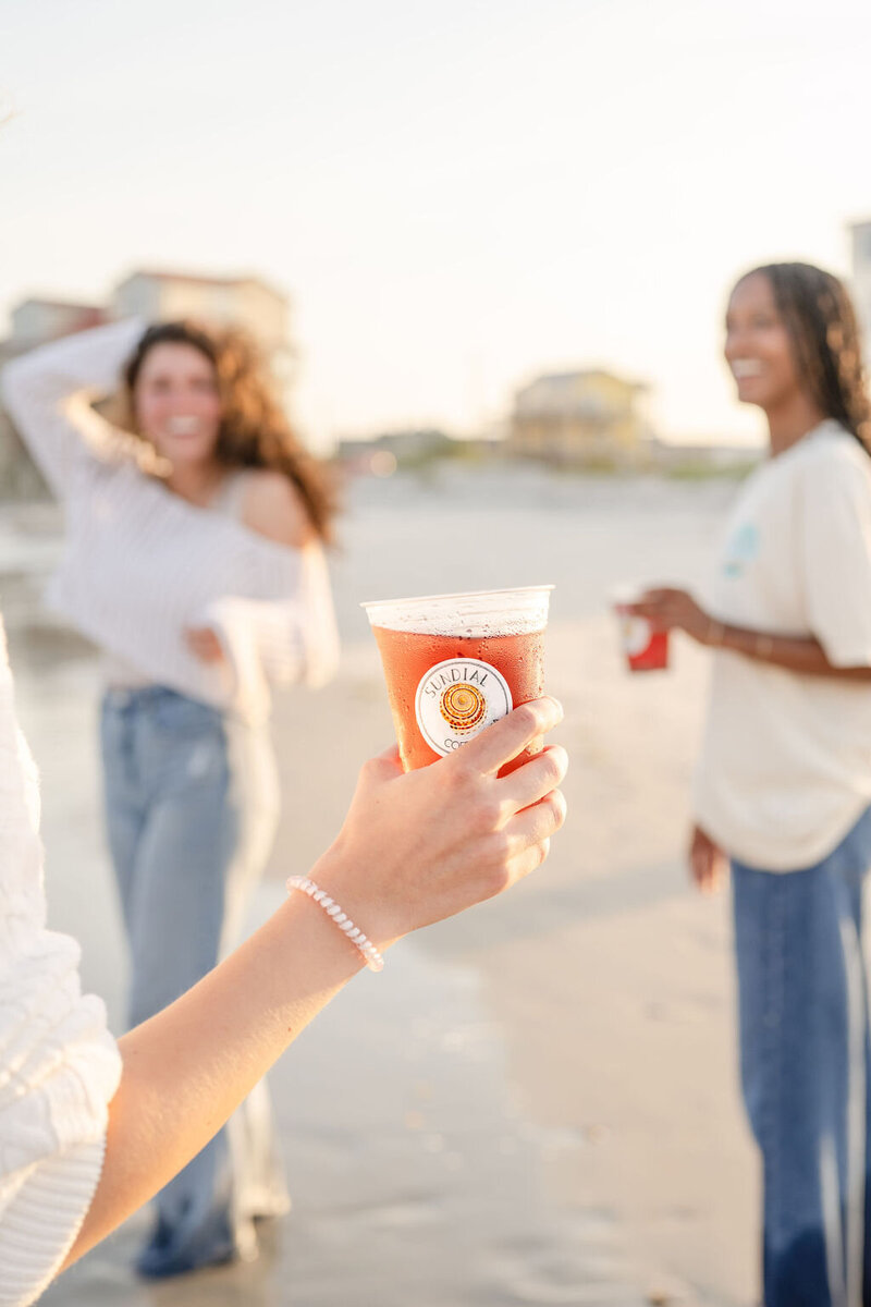 woman holds an iced hibiscus tea from sundial coffee and tea on the beach with two of her friends smiling in the background