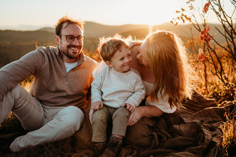 mom and toddler smile at each other during sunset on the mountain while dad looks on smiling