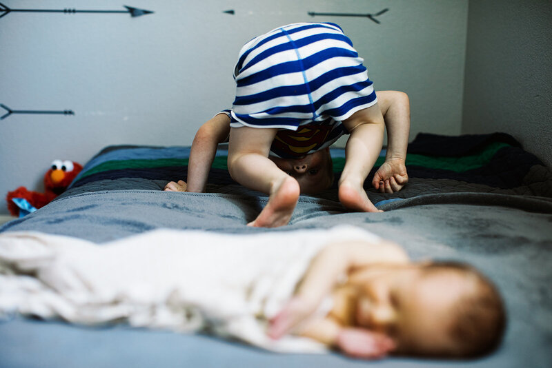 Playful toddler doing gymnastics on the bed next to his newborn brother during a relaxed in-home family session in Erie, Colorado