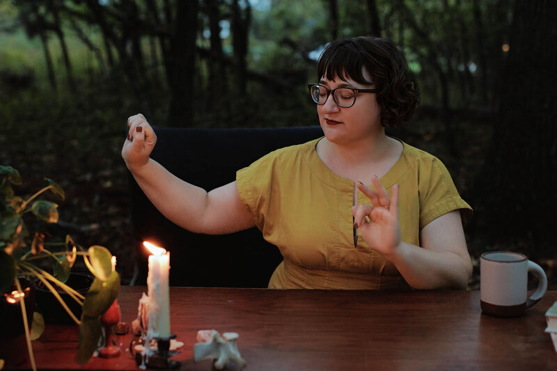 Ash Stinson sits at a wood table in the trees practicing energy work wearing a mustard yellow dress