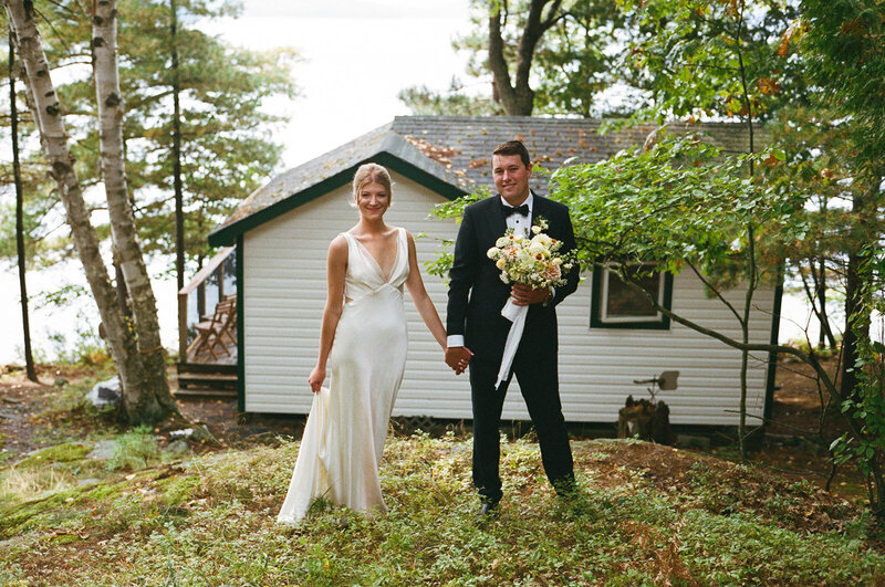A bride and groom stand holding hands outdoors, captured elegantly by a Toronto wedding photographer at a Muskoka cottage wedding. The bride is in a white dress with a bouquet, while the groom dons a suit. A small building and trees provide a picturesque backdrop to this beautiful moment.