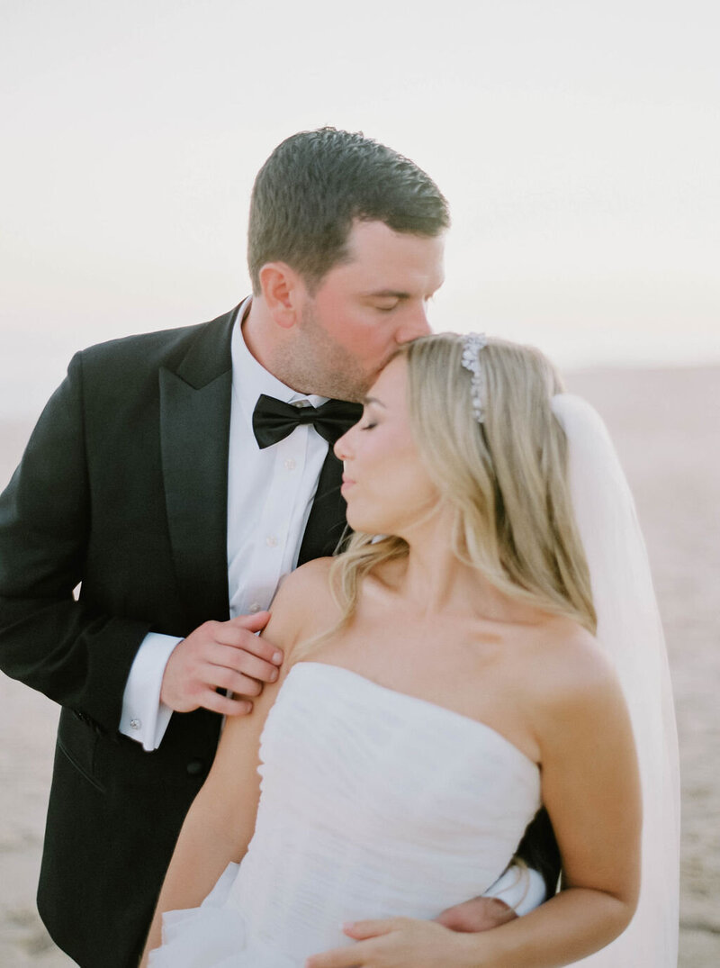 Rhode Island Wedding Photographers | A groom in a tuxedo kisses the bride's forehead on a beach. The bride wears a strapless dress and veil, eyes closed.