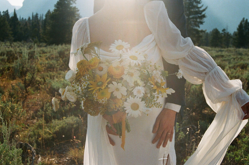 A couple stands on large boulders in the desert, surrounded by towering saguaro cacti, with one person helping the other balance atop the rocks.