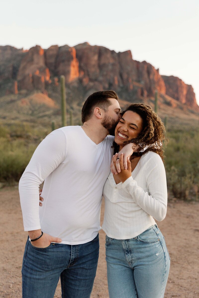 Boyfriend kisses his fiancé on the cheek while she giggles.
