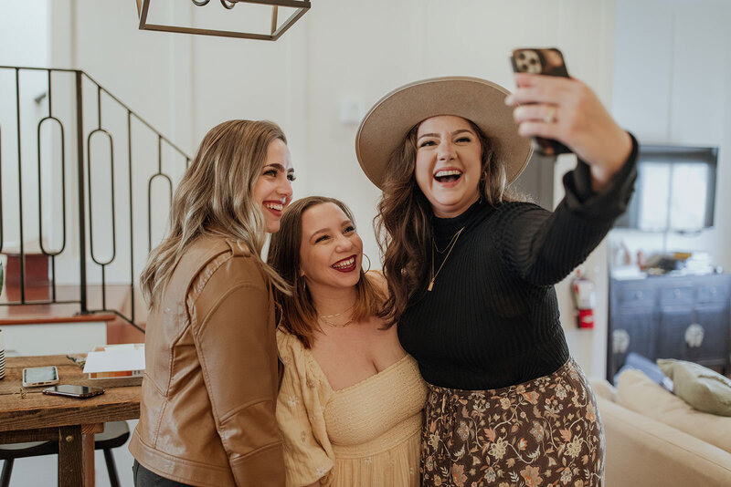 Three women smile and pose together for a selfie in a cozy, well-lit living room. Happy and cheerful, they look like friends celebrating after completing a white label web design course. One wears a wide-brimmed hat, another a brown jacket, and the third a cream dress.