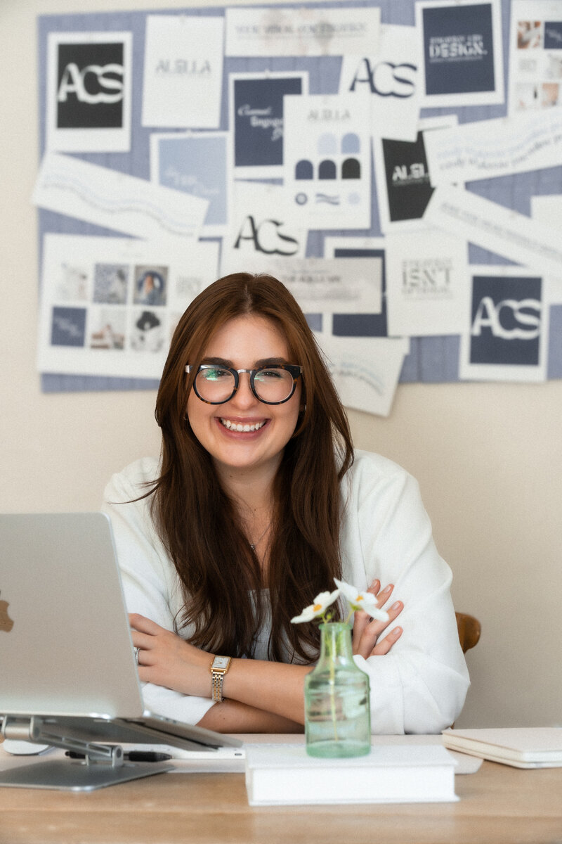 A smiling woman with dark hair and glasses, wearing a white blazer, looking directly at the camera while holding an open silver laptop. She appears professional and approachable, representing the comprehensive "Full Brand Experience" service.