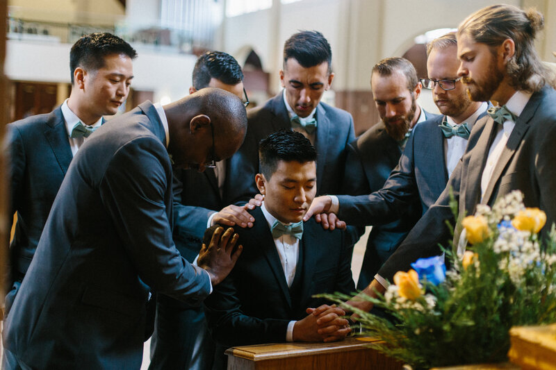 Groom kneeling down in church while groomsmen lay their hands on him