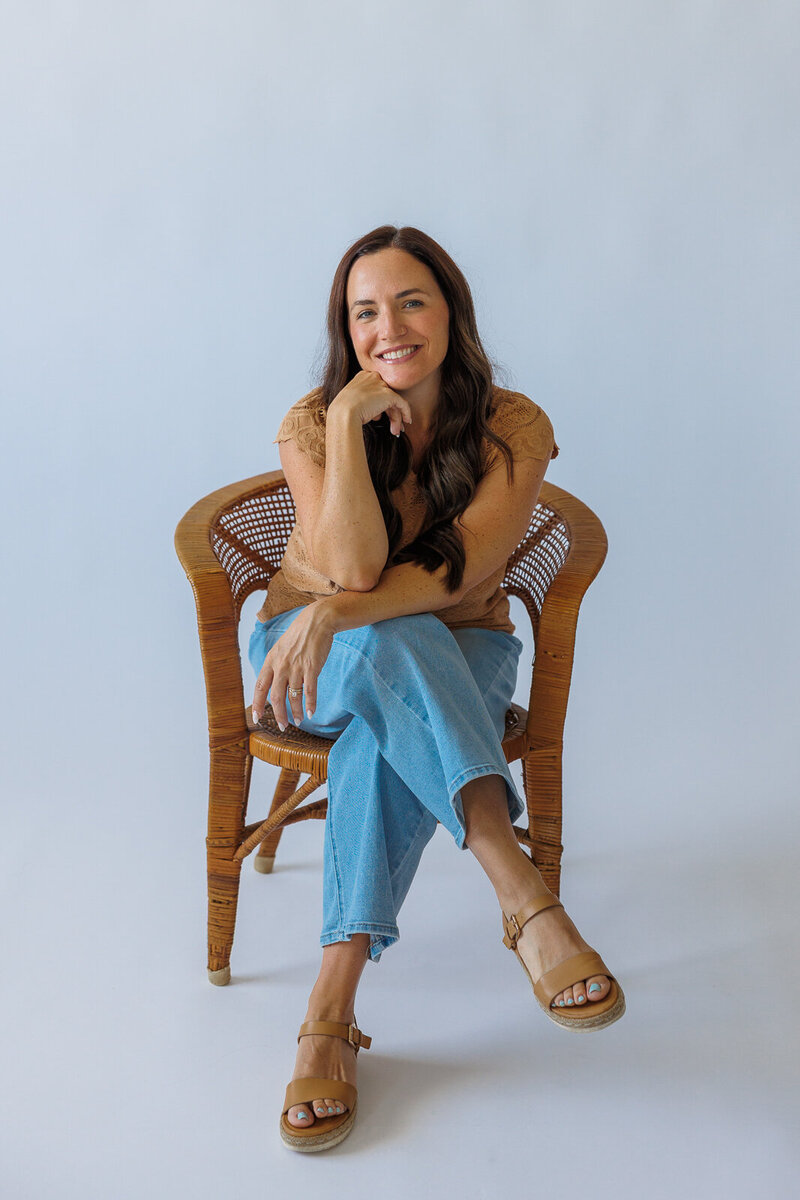 Stephanie Wilson sits in a rattan chair on a white photography backdrop while smiling at the camera