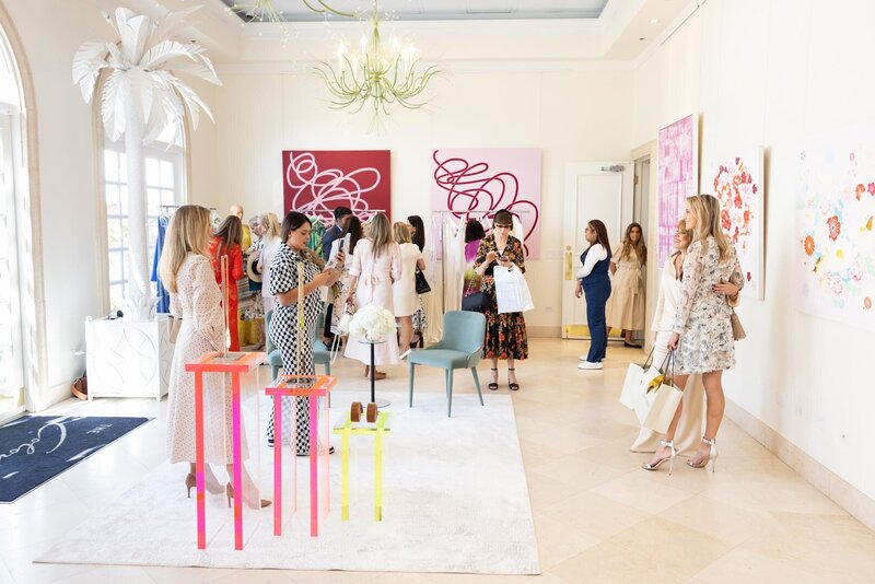 Guests mingle and take photos during the BCNY Palm Beach 2025 Luncheon, held in a bright, modern space with white décor, neon acrylic display tables, and bold abstract red artwork on the walls.