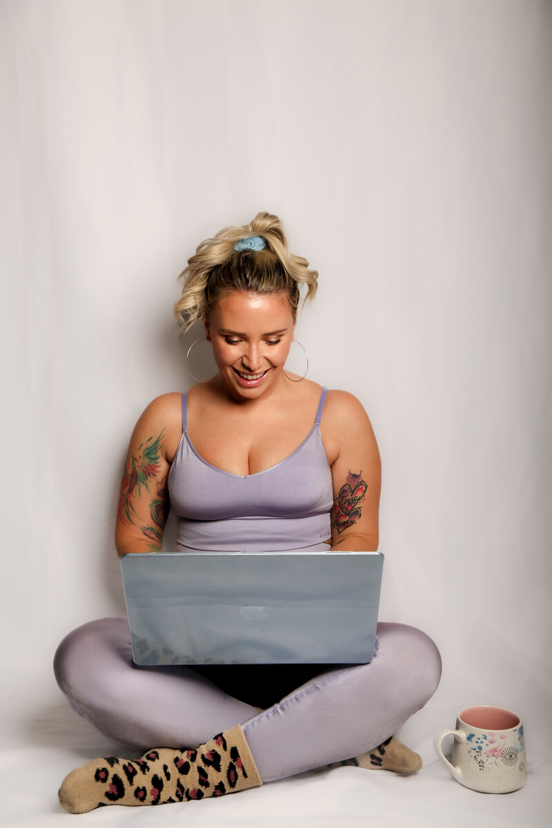 Robin is wearing a light purple athleisure set and is sitting on the floor with her laptop and a cup of coffee, creating courses to help mothers reclaim their energy and peace
