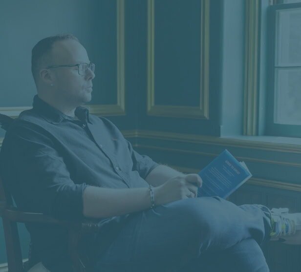 Dr. Mike Stokes sitting by a window reading a book, with a teal overlay effect.