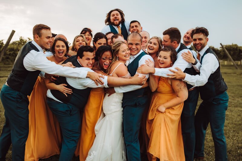 A bride and groom surrounded by their bridesmaids and groomsmen. Everyone is embracing them in a hug and smiling and laugjhing