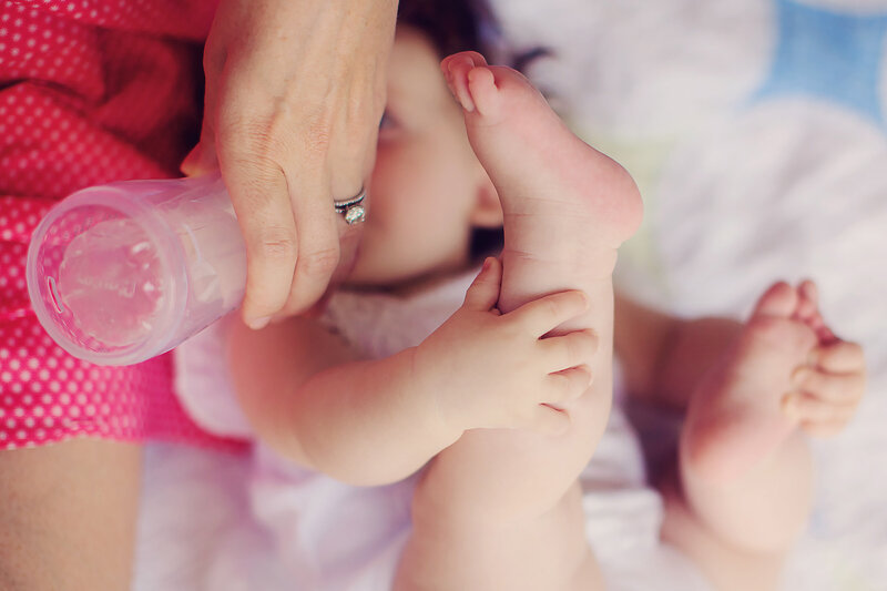 Baby ls laying on a quilt on her back, holding her feet, while her Mom feeds her a bottle.