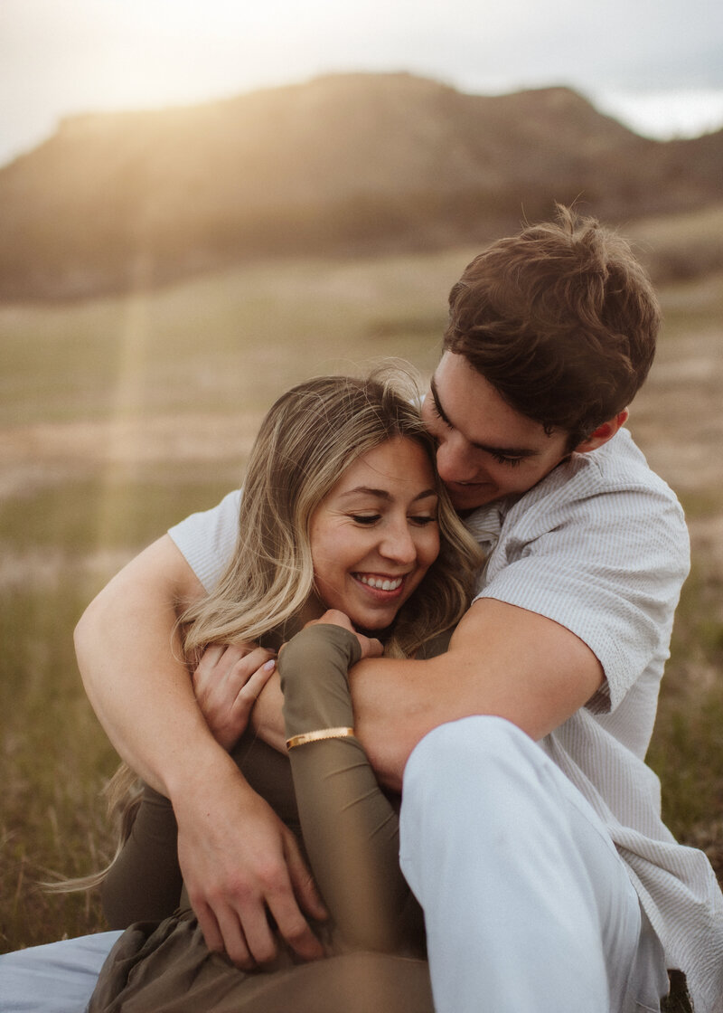 A young couple sit and hold each other in the golden hour glow at Dawson Butte Open Space Colorado. 
