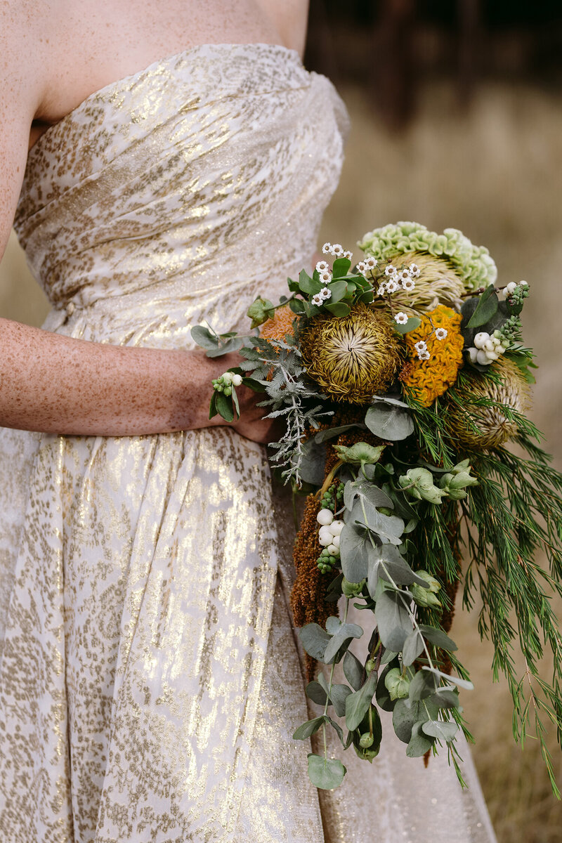 Bride holding a stunning bouquet of orange flowers and greenery on her wedding day, captured by Carry Your Heart Photography. The delicate arrangement complements her elegant lace gown, and the joy on her face reflects the happiness of the special day. This close-up shot highlights the intricate details of the bouquet and the bride’s radiant beauty, set against the charming backdrop of the wedding venue.