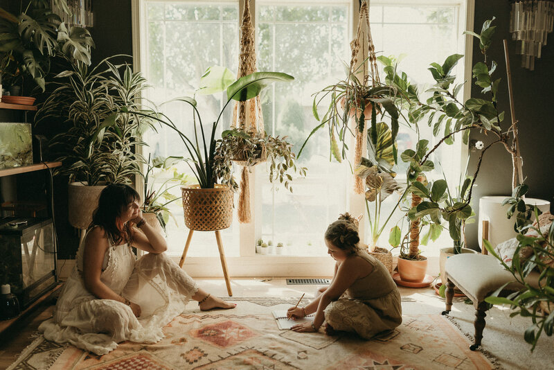 In-home lifestyle portrait of Mom looking at daughter drawing. Many indoor plants in boho home.