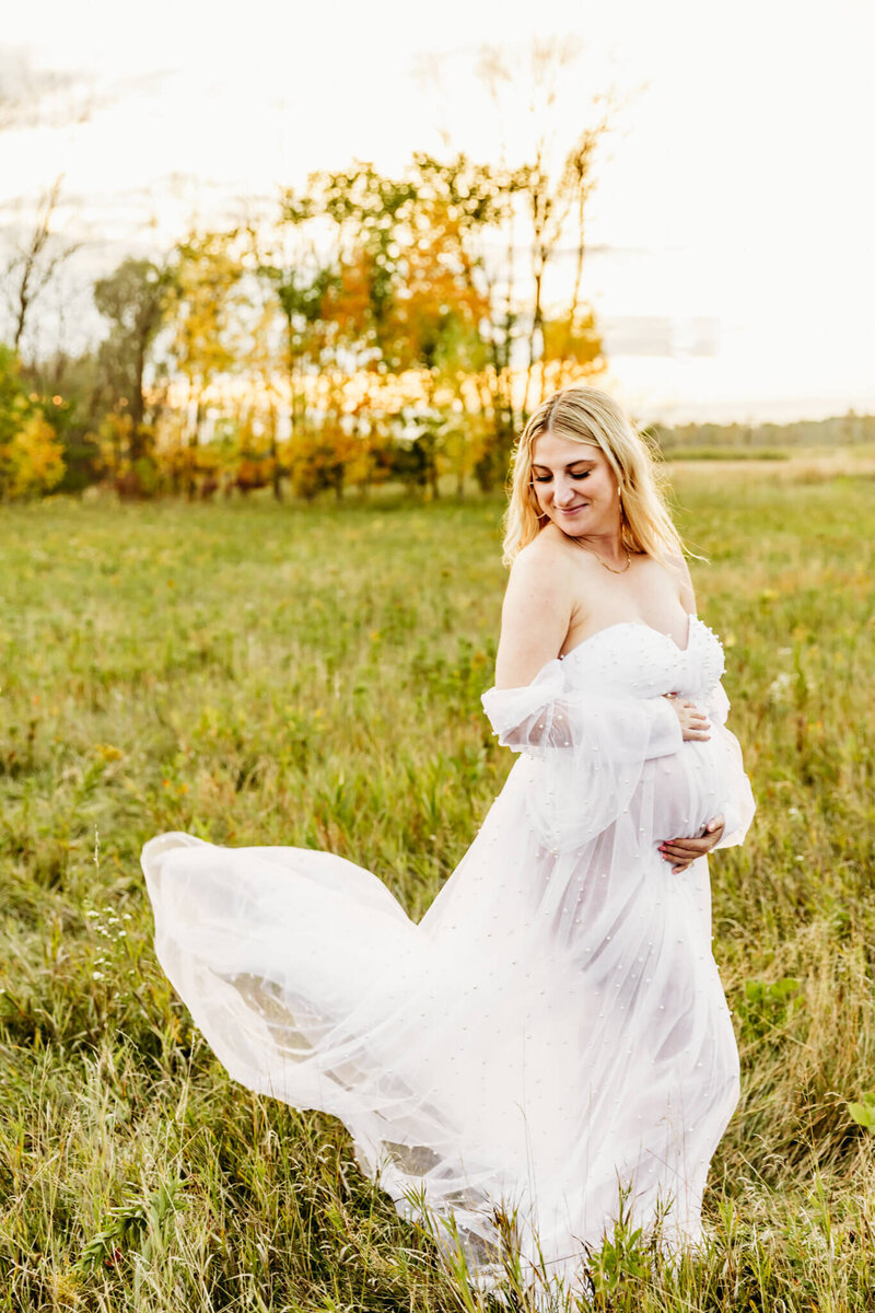 beautiful pregnant woman in a white gown looking at shoulder as she embraces her baby bump and dress is flying