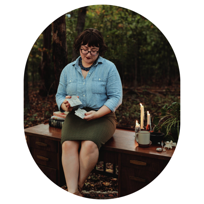 Ash sits on a wood table nestled amongst trees in the forest and shuffles the cards of the Energy Archaeology Oracle Deck