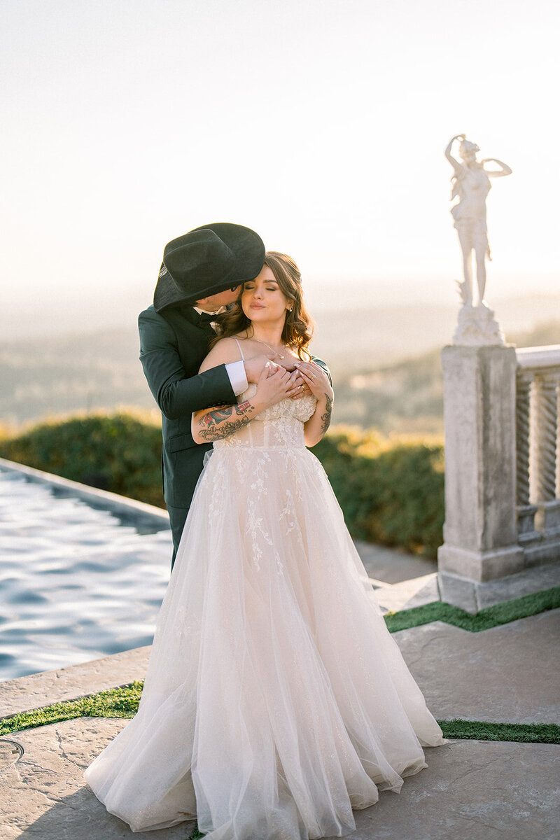 This romantic California wedding moment captures the couple in a tender embrace, bathed in the golden light of sunset by the water. The bride, in her ethereal gown, leans into her groom as he wraps his arms around her, creating an intimate and serene scene. The picturesque backdrop of rolling hills and statues adds to the fairytale ambiance, perfectly blending elegance with the natural beauty of a California wedding.
